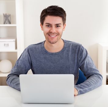 Smiling handsome casual young man sitting at a table using a laptop in a home office