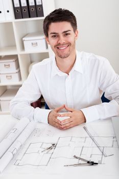 Confident young architect sitting at his desk in his office with a set of rolled blueprints and a hardhat smiling at the camera