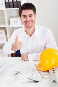 Confident young architect sitting at his desk in his office with a set of rolled blueprints and a hardhat smiling at the camera