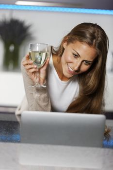 Young woman in her kitchen using a laptop while enjoying a glass of wine.