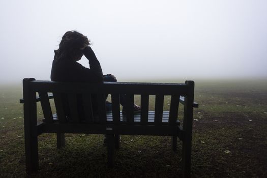 Woman seated on bench ponders over the misty field pending future days.