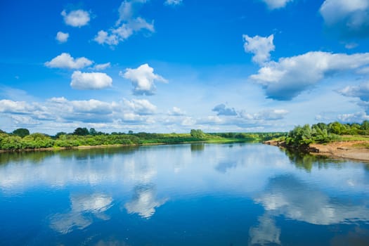 Wide river with reflection and green bush on coasts and blue cloudy sky