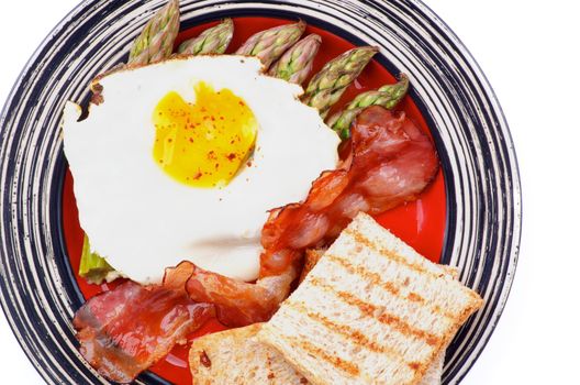 Tasty Breakfast with Fried Egg Sunny Side Up on Delicious Asparagus Sprouts, Roasted Bacon and Toasts on Striped Plate closeup on white background. Top View