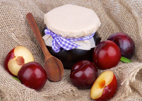 Arrangement of Jar of Plum Jam with Wooden Spoon,  Ripe Purple Plums Full Body and Halves closeup on Burlap background