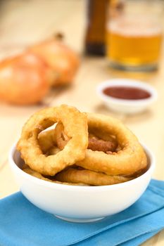 Freshly prepared homemade beer-battered onion rings in a bowl with beer, ketchup and onions in the back (Selective Focus, Focus on the front of the onion ring on the right)