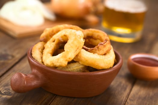 Freshly prepared homemade beer-battered onion rings in a rustic bowl with ketchup on the side (Selective Focus, Focus on the front of the onion ring on the right)