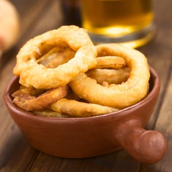 Freshly prepared homemade beer-battered onion rings in a rustic bowl with beer in the back (Selective Focus, Focus on the front of the onion rings on top)