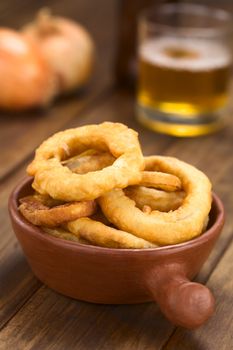 Freshly prepared homemade beer-battered onion rings in a rustic bowl with beer in the back (Selective Focus, Focus on the front of the onion rings on top)