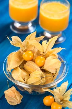 Physalis berry fruits (lat. Physalis peruviana) with husk in glass bowl with fresh physalis juice in the back on blue wood (Selective Focus, Focus on the open physalis berries in the bowl)