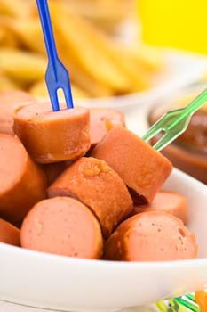 Fried sausage pieces with two small plastic forks, French fries and ketchup in the back (Selective Focus, Focus on the sausage on the top) 