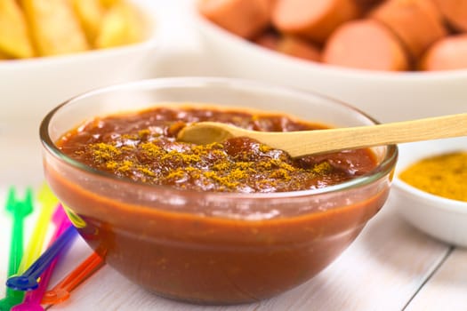 Curry ketchup sauce made to dip fried sausages and French fries served in glass bowl with wooden spoon, colorful plastic party forks on the side, fried sausage pieces and French fries in the back (Selective Focus, Focus one third into the sauce)  