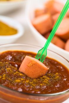 Fried sausage on plastic party fork dipped into curry ketchup sauce, fried sausage pieces and curry powder in the back (Selective Focus, Focus on the sausage piece on the sauce)  