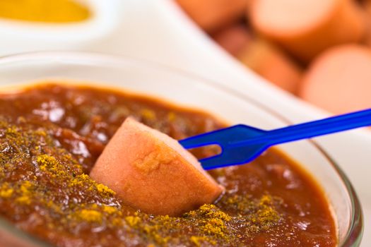 Fried sausage on plastic party fork dipped into curry ketchup sauce, fried sausage pieces and curry powder in the back (Selective Focus, Focus on the sausage piece on the sauce)  