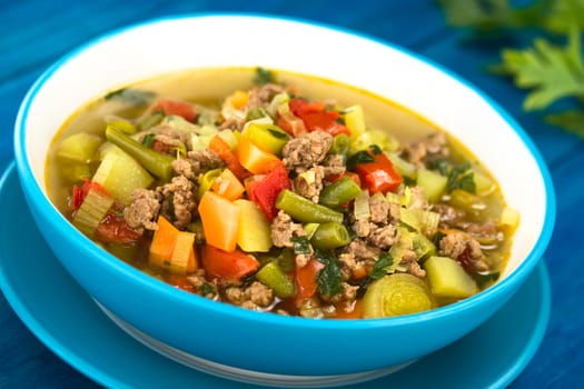 Vegetable soup with mincemeat, green bean, potato, leek, carrot, tomato and parsley served in blue bowl on blue wood (Selective Focus, Focus one third into the soup) 