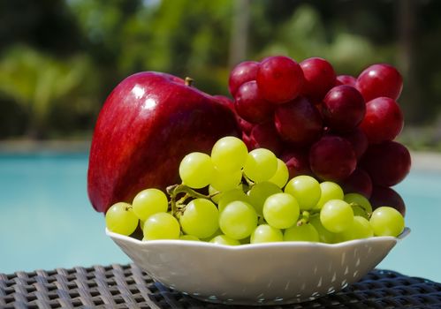 Red apple and grape in white plate on table by the swimming pool