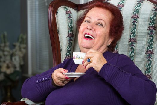 Senior Woman having good time while having her Turkish Coffee, she is wearing a purple sweater and sitting on comfortable classic big seat