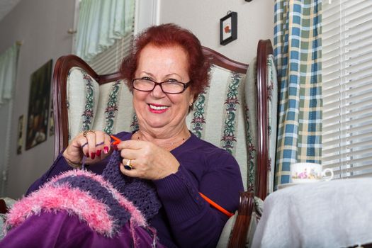 Red haired senior lady looking at you right abow her glasses while knitting her purple scarf