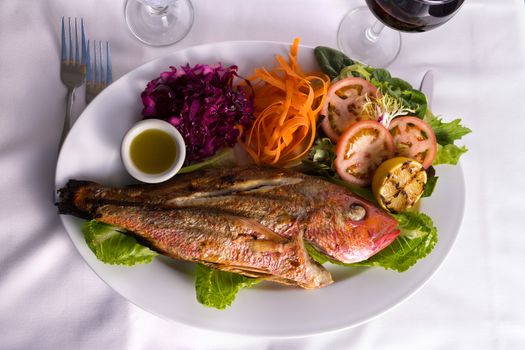 Full cooked tilapia fish served in a plate with vegetables and fish sauce complimented with red vine