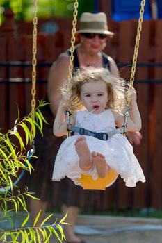Toddler girl having fun on the swing while having candy with her bare naked feet and pushed by her grandmother with the hat