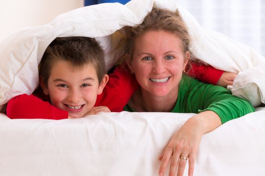 Mother and her son under the blanket with Christmas color pajamas, smiling to you