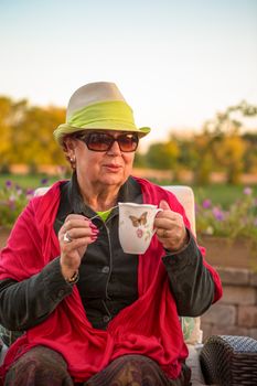 Senior woman with a straw green hat, sitting at the patio and showing her hot tea, perhaps she wants to stay warm