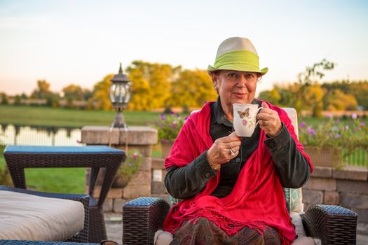 Senior woman with a straw green hat, sitting at the patio and showing her hot tea, perhaps she wants to stay warm