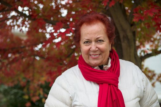 Red hair senior lady looking at you happily and trustfully with her red scarf and white coat under the tree with red falling leaves