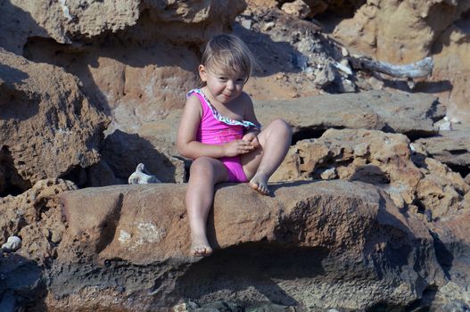 Two-year-old girl in a pink bathing suit sitting on stones and smiling                               