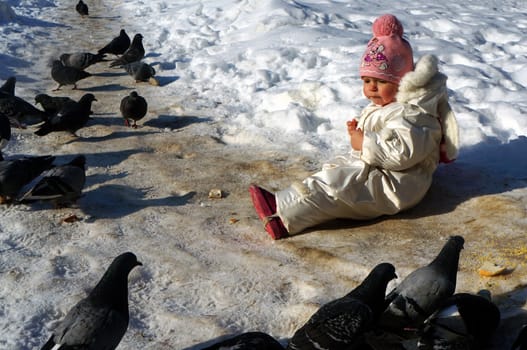 Two-year-old girl in white kombnezone sitting in the snow and feeding pigeons                               