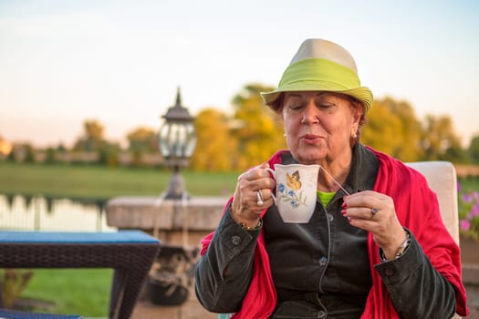 Senior woman with a straw green hat, sitting at the patio and blowing her hot tea, perhaps she wants to stay warm