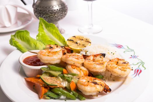 Shrimp skewers are grilled and garnished with veggies and rice served on a white plate along with a silver pitcher