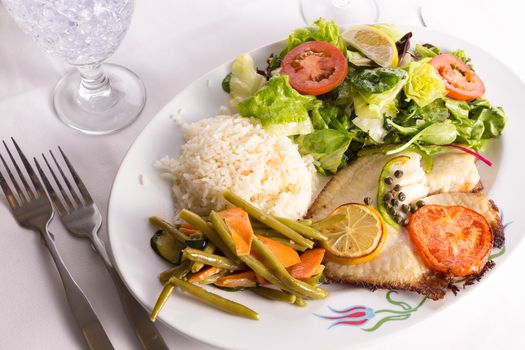 Tilapia fish cooked with capers, lemon and tomatoes and served in a plate with vegetables and pilaf rice