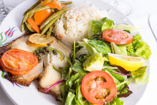 Tilapia fish cooked with capers, lemon and tomatoes and served in a plate with vegetables and pilaf rice