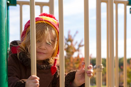 Little girl at the playground looking at you mischievously behind the yellow bars with her red winter hat