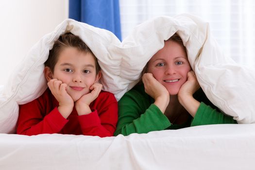 Mother and her son under the blanket with Christmas color pajamas, smiling to you