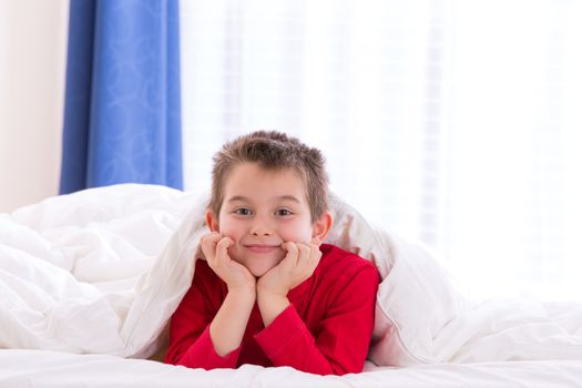 Close up portrait of a eight years old boy laying on the bed on looking at  you in his bedroom at home. He looks calm and happy