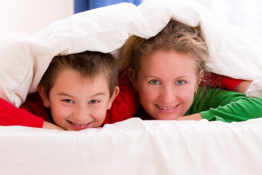 Mother and her son under the blanket with Christmas color pajamas, smiling to you cheerfully
