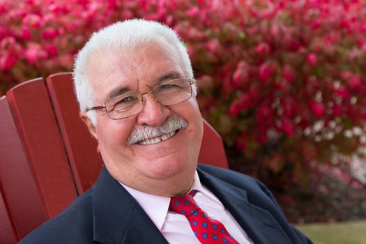 White hair senior businessman outside on a red chair smiling large at you