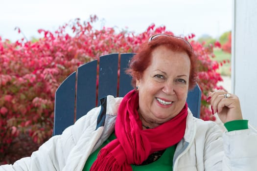 Red hair senior woman outside on a blue chair smiling large with her Christmas color clothing