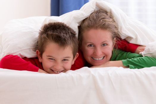 Mother and her son under the blanket with Christmas color pajamas, smiling to you while having great moments