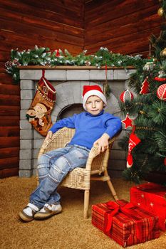 Boy in hat sitting in wicker chair near Christmas tree and gifts