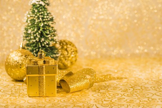 Golden Christmas ornaments, golden Christmas decoration in present box shaped, golden shiny ribbon and snowed Christmas tree on golden background