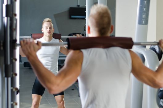 Attractive young man in gym mirror doing squats with equipment