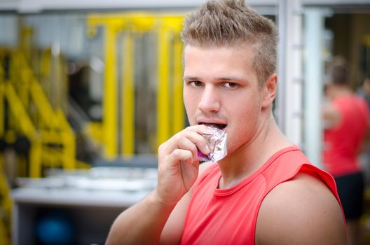 Handsome young man in gym eating cereal bar, looking at camera