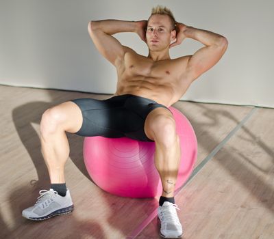 Shirtless muscular young man training, exercising abs on fitness ball