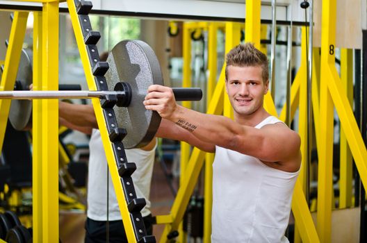 Smiling handsome young man in gym putting weight disck on barbell, looking at camera