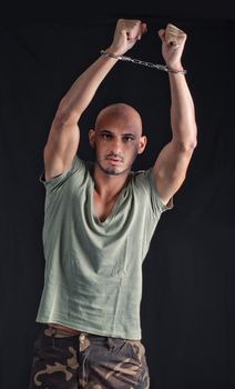Bald young man with handcuffs, holding arms up in the air, on dark background