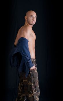 Bald young man standing, taking off sweatshirt, wearing military pants