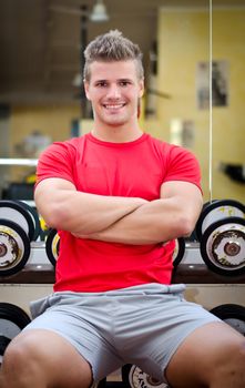 Handsome young man in gym sitting on dumbbells rack, smiling and looking in camera