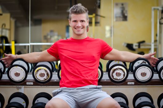 Handsome young man in gym sitting on dumbbells rack, smiling and looking in camera